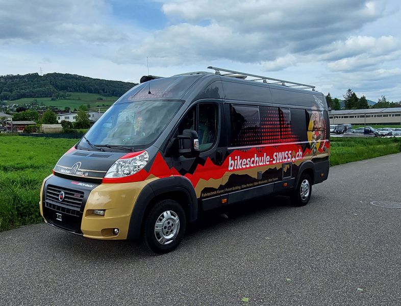 Transportbus mit seitlicher Beschriftung Bikeschule Swiss und Bergpanoramamotiv.