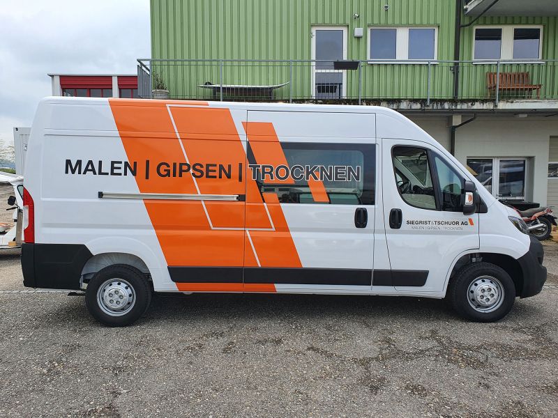 Transportbus weiss mit seitlicher Aufschrift Siegrist & Tschuor AG, mit orangen Streifen.